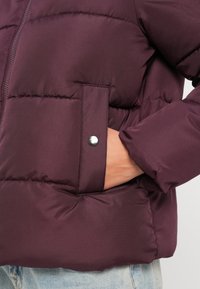 Chaqueta acolchada burdeos con diseño acolchado, cierre de cremallera, bolsillo lateral con broche de metal y textura suave y lisa.