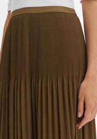 Jupe plissée vert olive avec une taille ajustée, ornée de plis verticaux et horizontaux, confectionnée dans un tissu doux pour un design fluide.