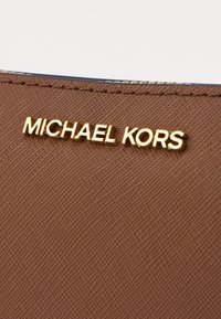 Brūns teksturēts materiāls ar metālisku zelta "MICHAEL KORS" logo lielajiem burtiem. Virsma ir ar smalku austā rakstu.