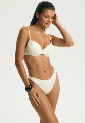 Žena v bielom bikinách s čiernym náramkom a náušnicami pózuje na svetlom jednofarebnom pozadí, ruka sa dotýka hlavy, pozerá dole.