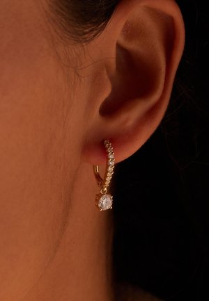 Boucle d'oreille créole en or avec de petites pierres transparentes le long du cercle et une plus grosse pierre précieuse claire pendante sur une oreille percée.
