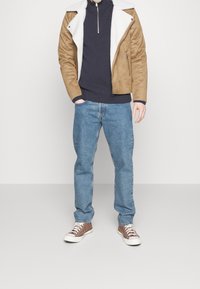 Brązowa kurtka z imitacji zamszu z białym kołnierzem z baranka, granatowa bluzka z długim rękawem, jasnoniebieskie dżinsy oraz brązowe sneakersy z białą podeszwą.