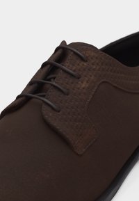 Καφέ σκούρο δερμάτινο επίσημο παπούτσι με υφαντή υφή στην περιοχή των κορδονιών και μαύρα κορδόνια, κοντινή λήψη στο μπροστινό και πλευρικό μέρος.