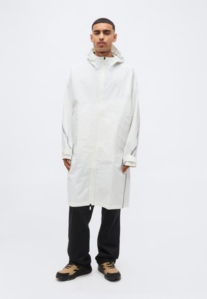 Άνδρας που φοράει μακρύ λευκό αδιάβροχο με κουκούλα, μαύρο παντελόνι και καφέ παπούτσια πεζοπορίας, στέκεται μπροστά σε λευκό φόντο.