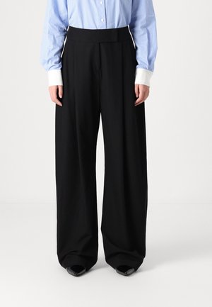 Persona che indossa pantaloni neri a gamba larga, camicia a righe blu e bianche con polsini bianchi e scarpe nere con punta appuntita, in piedi su sfondo bianco.