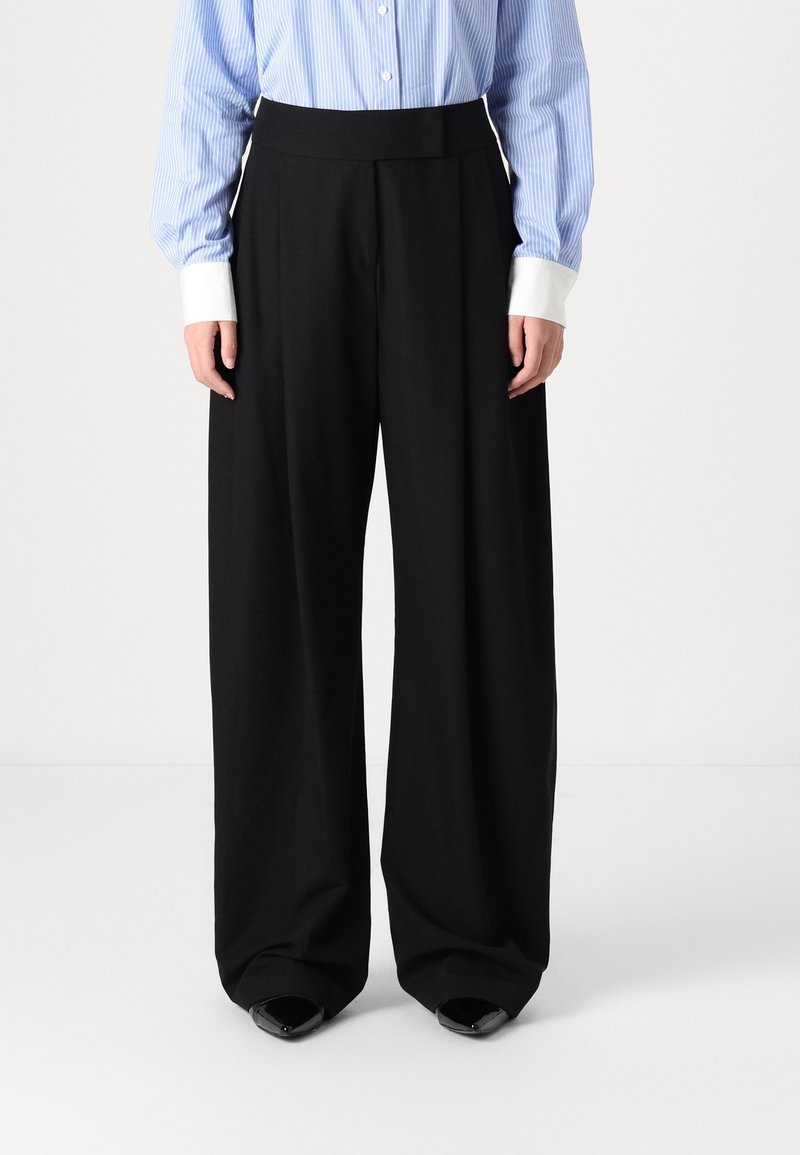 Personne portant un pantalon noir à larges jambes, une chemise rayée bleu et blanc avec des poignets blancs, et des chaussures noires à bouts pointus, debout sur un fond blanc.