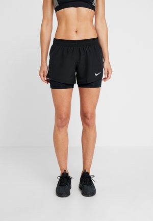 Kobieta ubraną w czarne, warstwowe szorty Nike, czarny stanik sportowy i czarne buty do biegania, stojąca na białym tle.