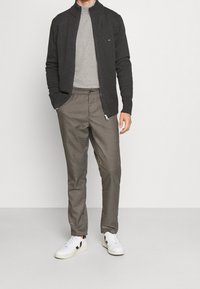 Grauer Reißverschlusscardigan über einem hellgrauen T-Shirt, kombiniert mit braun-weißen karierten Hosen und weißen Turnschuhen.