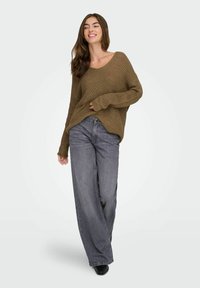 Maglione marrone oversize a maglia con scollatura a V e maniche lunghe, abbinato a jeans a gamba larga grigi e stivaletti neri, su sfondo bianco.