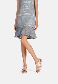 Abito in pizzo grigio con orlo a campana, caratterizzato da strisce orizzontali. Sandali beige con fasce incrociate e suola imbottita.