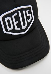 Gorra negra con un diseño estructurado, que presenta un prominente logotipo bordado en blanco "DEUS" en la parte frontal. Tejido suave con visera curvada.