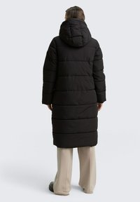 Zwarte pufferjas met een capuchon, gestikte secties voor isolatie en een lange silhouet, gecombineerd met lichtgekleurde broek en schoenen.