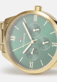 Orologio dorato con un quadrante verde e testurizzato. Presenta tre sottoquadranti, numeri romani e un bracciale in metallo dorato. Logo del marchio visibile.