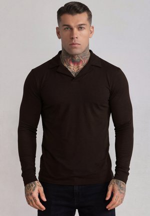 Man met tattoos op nek en handen, draagt een nauwsluitend zwart shirt met lange mouwen, staand tegen een effen lichte achtergrond.