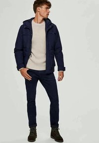 Mornarska podložena jakna s kapuco, v kombinaciji s svetlo kremo beljenim puloverjem z rebrastim vzorcem in temno modrimi hlačami. Obleka vključuje detajle z zadrgo in žepi.