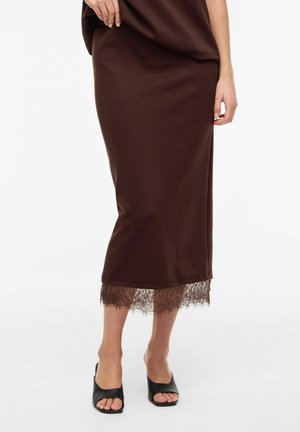 Mujer con una falda midi marrón con encaje en el dobladillo y mules negros de tacón abiertos, posando frente a un fondo blanco.