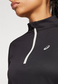 Crni sportski top s četvrtinim zipom, s bijelim detaljem na zipu i brendiranim logotipom. Izrađen od teksturiranog, rastezljivog materijala.