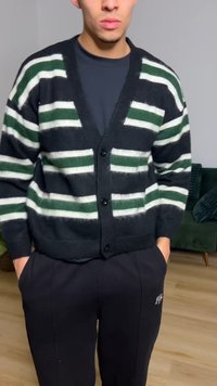 Schwarzer Cardigan mit grünen und weißen horizontalen Streifen, Knopfleiste vorne, gerippte Bündchen und Saum, getragen über einem dunklen Hemd und schwarzen Hosen.
