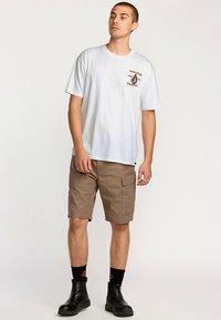 White short-sleeve T-shirt with "VOLCOM" logo on the chest, paired with tan cargo shorts; black boots and socks complete the outfit.