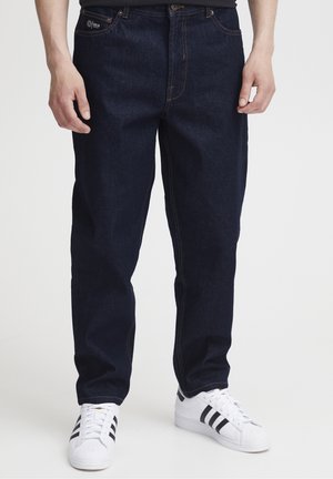 Mann trägt dunkle, gerade geschnittene Jeans und weiße Sneaker mit schwarzen Streifen, steht vor einem schlichten hellen Hintergrund.