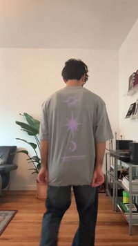 Mand med mørkt hår iført en grå t-shirt med lilla himmellegemer, stående i et stue med planter og hylder.