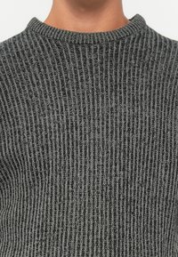 Primo piano di una persona che indossa un maglione a coste grigio scuro con collo rotondo su uno sfondo chiaro e uniforme.