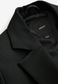Sort blazer med høj hals, tekstureret stof og glat inderside. Har et mærketikette, der viser "NEXT" og plejeinformation.