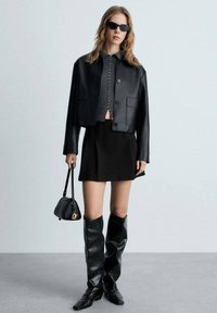 Giacca cropped nera in pelle, top grigio a maglia attillato, mini gonna plissettata nera, stivali neri al ginocchio e piccola borsa nera.