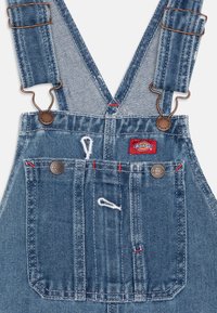Jeansoverall med justerbare stropper, metallbeslag, frontlomme, røde sømdetaljer og en Dickies-logo-patch på brystet.