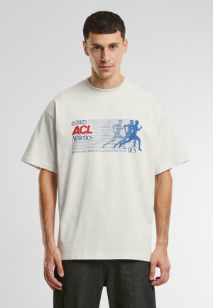 T-shirt oversize gris clair avec un design graphique bleu représentant des figures athlétiques et du texte, fabriqué en tissu de coton doux. Manches courtes.