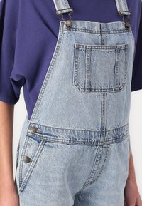 Salopettes en denim bleu clair avec bretelles réglables, une poche sur la poitrine et des accents en métal contrastés.
