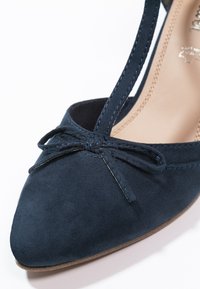 Chaussure en daim bleu marine avec un bout pointu, une bride aux chevilles et un délicat nœud à l'avant. Texture lisse avec des coutures contrastées.