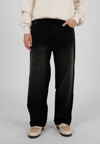 Jeans in denim nero con vestibilità rilassata, leggermente scoloriti, cinque tasche e una cintura robusta. Abbinati a calzature color crema.