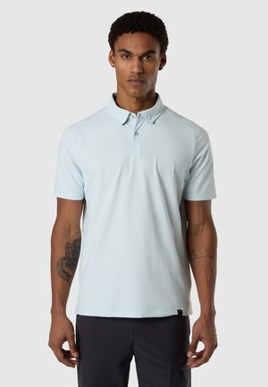Ein Mann steht frontal, trägt ein hellblaues Polohemd mit kurzen Ärmeln und dunkle Hosen, mit sichtbaren Tätowierungen auf beiden Unterarmen.
