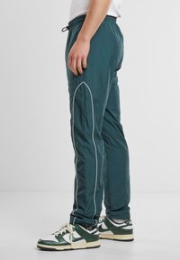 Pantalones ligeros en color aqua con ribetes laterales en blanco, cinturilla elástica y un ajuste relajado, combinados con zapatillas verdes y blancas. Textura suave.