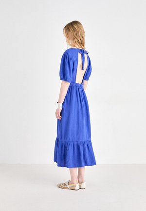 Vrouw staat in een blauwe midi-jurk met pofmouwen, een open rug die in de nek is gestrikt, witte gestreepte ballerina's en een zilveren manchetarmband.