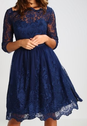 Kvinde iført knælange navyblå blondekjole med 3/4-ærmer og indviklede blomster mønstre, hænder forsigtigt foldet ved taljen.