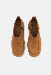 Brune semskede slip-on sko med rund tå, elastiske sidede accenter og en syet design; har en teksturert gummisåle og en stropp.
