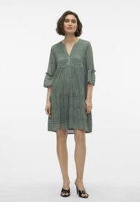 Robe en dentelle vert menthe avec un décolleté en V, des manches 3/4 et des poignets à volants, présentant des motifs complexes et un ourlet à plusieurs niveaux.