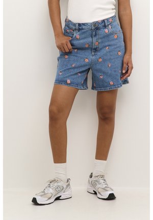 Person iført blå denimshorts, der går til midt på låret, med pink blomsterbroderi, hvide sokker, hvide New Balance-sneakers og en hvid top.