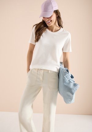 Lächelnde Frau in beigen Hosen und weißem T-Shirt mit Streifen hält eine hellblaue Jacke, trägt eine rosa Baseballkappe und steht vor einem hellen Hintergrund.