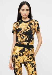 Γυναίκα που φοράει μαύρο και χρυσό μπλουζάκι με μανίκια μέχρι τον αγκώνα και παντελόνι σε μπαρόκ σχέδιο, με ζώνη μέσης "Versace Jeans Couture".