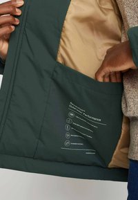 Dark green waterproof jacket interior with a gold lining, featuring a side pocket and printed care instructions. Smooth texture.
