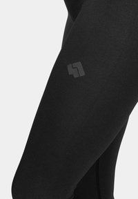 Czarne legginsy z elastycznego materiału charakteryzują się gładką fakturą i małym, subtelnym nadrukiem logo z boku.