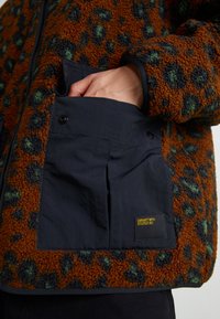 Fleecejakke med brune, grønne og sorte leopardpletter; har en knaplukket klaplomme og tekstureret stof langs kanterne.