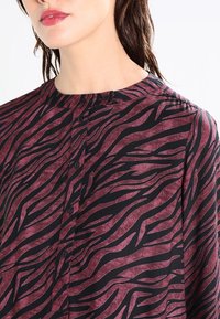 Blouse à manches longues avec col haut, motif rayé tigre bordeaux et noir, avec une fermeture à boutons sur le devant et une texture de tissu douce.