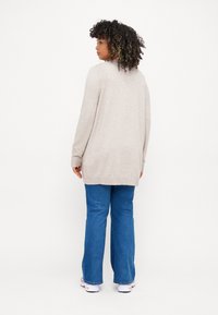Beige long cardigan with ribbed cuffs and hem, paired with blue wide-leg jeans and white sneakers, seen from the back against a white background.
