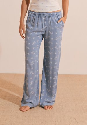 Persoon draagt lichtblauwe losse pyjamabroek met witte croissant- en tekstpatronen, staat op blote voeten op een beige tapijtvloer.