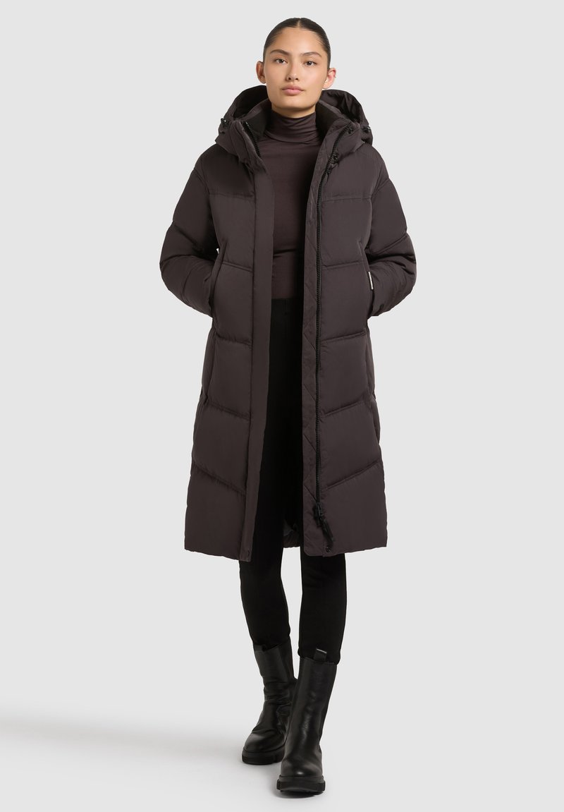 Abrigo puffer largo y oscuro con capucha, que cuenta con cierre de cremallera, bolsillos laterales y un diseño acolchado. Llevado sobre un suéter de cuello alto y pantalones negros.