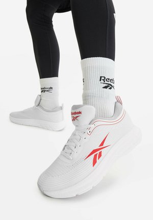 Persona che indossa sneakers bianche Reebok con logo rosso e calzini bianchi Reebok, vestita con leggings neri, in piedi su una superficie bianca.